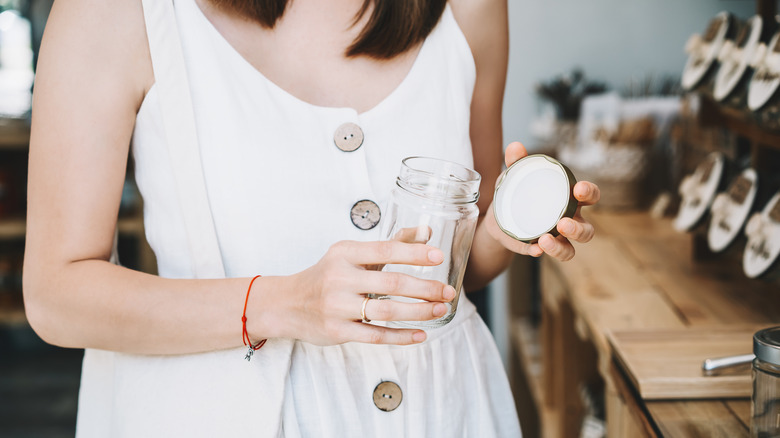 A woman standing in a kitchen holds an empty glass jar in one hand and the metal lid in the other.