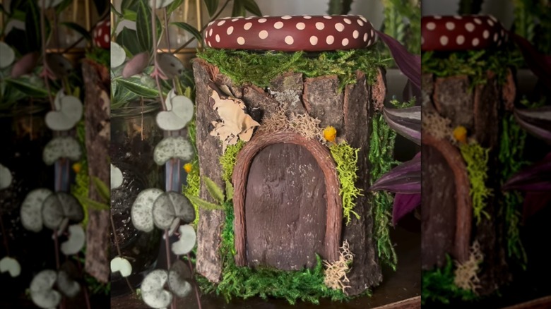 A circular fairy house topped with a dark red cap with white dots is decorated with tree bark and preserved moss