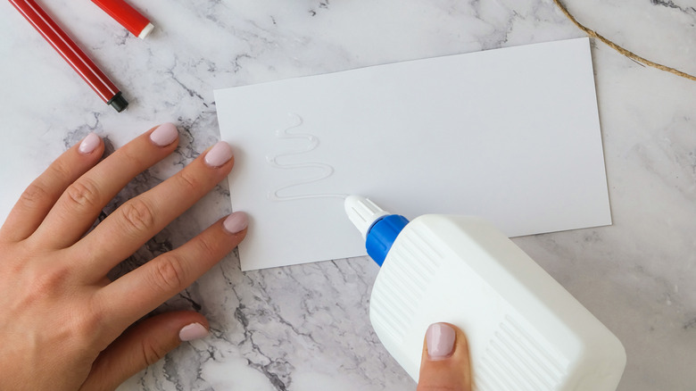A hand with painted pink nails holding a white bottle of school glue and squirting it onto a piece of white paper