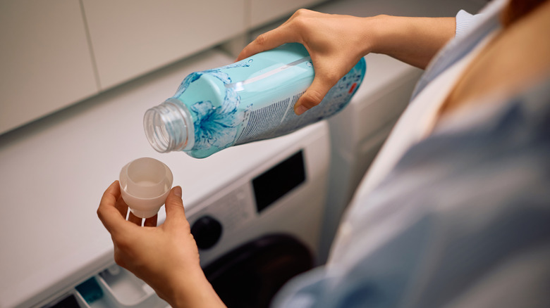 A person pours fabric softener into a bottle cap before above a washing machine.