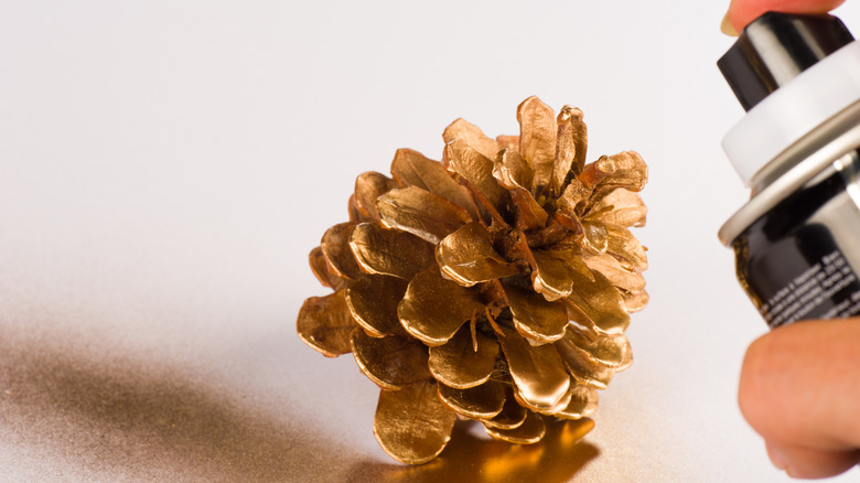 A hand holds a can of spray paint while painting a small pinecone gold.