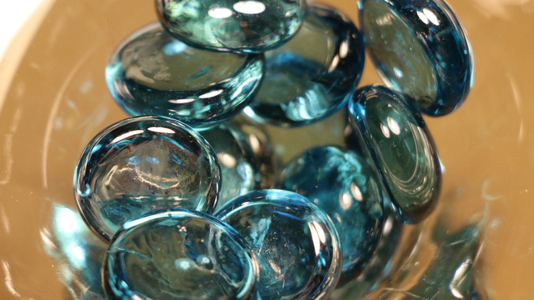 Teal glass circular stones piled into a glass container that sits on top of a light brown surface.