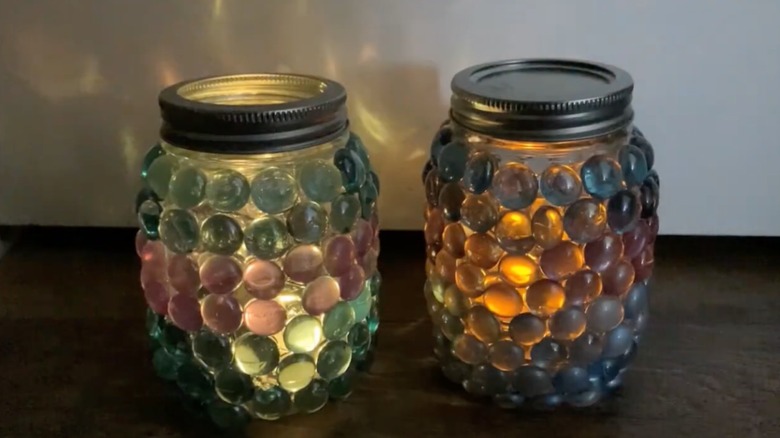 Two Mason Jars, covered with blue and purple glass gemstones, are lit with tealights.