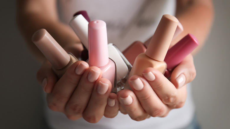 A pair of hands with nude-colored fingernails are holding nail polish bottles in shades of white, nude, and pinks.