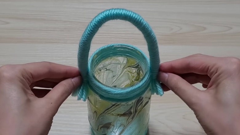 Two hands are holding an arched handle made of teal yarn over a yellow and gray marbled glass jar.