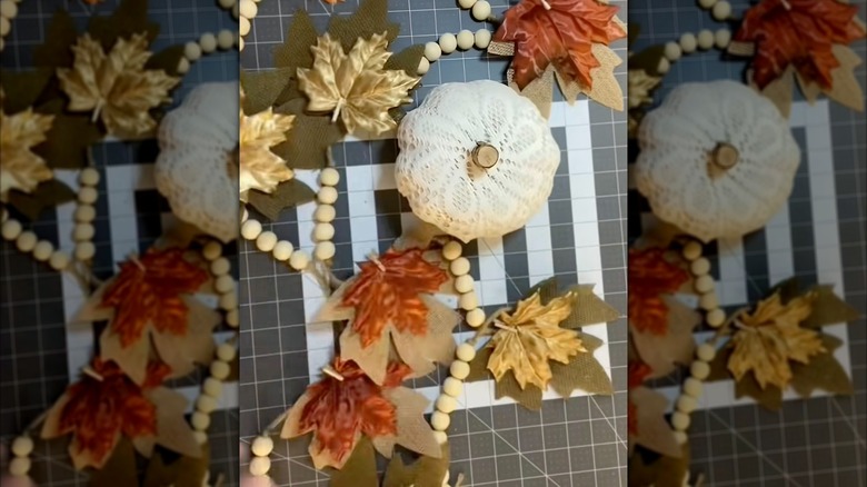DIY fall decor on a table, featuring a foam pumpkin wrapped in a doily surrounded by artificial fall flowers and beads on a string
