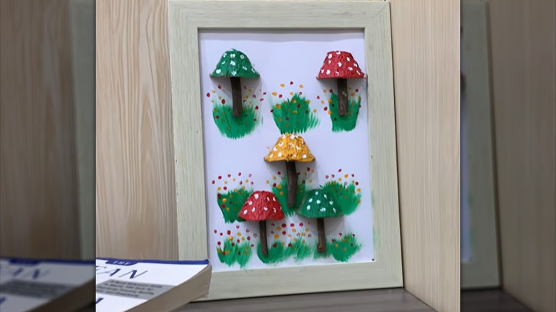 A framed white canvas decorated with mushrooms made from painted egg cups and sticks