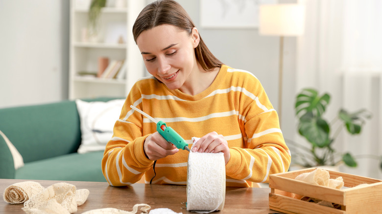 A woman in a striped sweater hot gluing white lace fabric onto a cylinder-shaped object