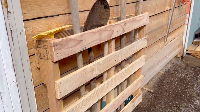 A wood pallet mounted to a wooden wall with tools like a shovel standing up within it