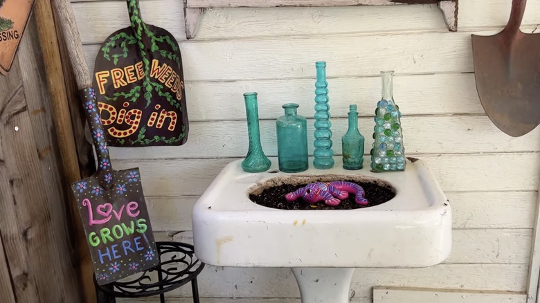 Shovel signs with phrases painted on their scoops propped next to an old kitchen sink filled with soil and fake worms and adorned with vintage glass bottles.