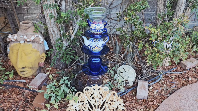 DIY bird bath made from teapots, blue plates, and a clear bowl on top in a garden surrounded by plants