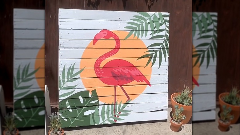 An old wood pallet repurposed into a painting with a flamingo, sun, and green plants painted on it.