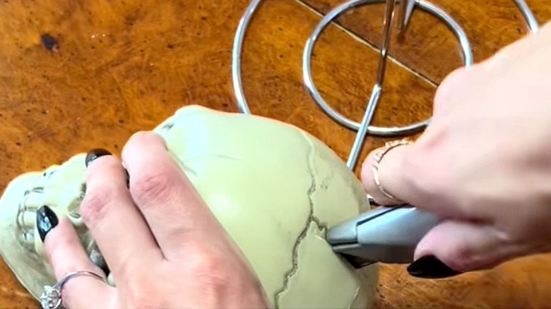 Close-up of a person using a box cutter to cut into a prop skull with the bottom of a paper towel holder seen in the background