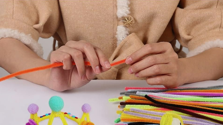 A woman bends an orange pipe cleaner she is holding in two hands above a table with more pipe cleaners and craft items on it.