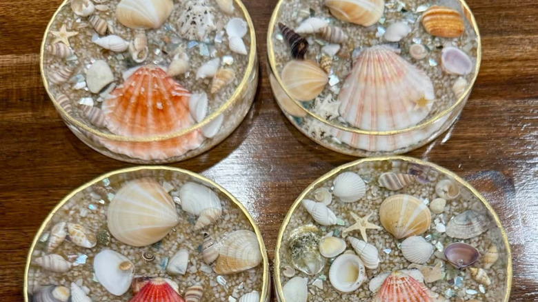 Four round coasters made with clear resin, sand, and seashells set on a wooden tabletop.
