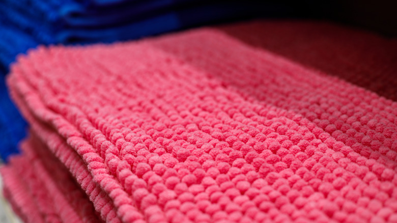 A folded pink fluffy bath mat with some blue towels in the background.