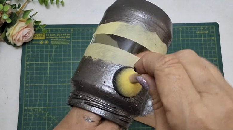 Person dabbing metallic paint on a glass jar with masking tape strips on it