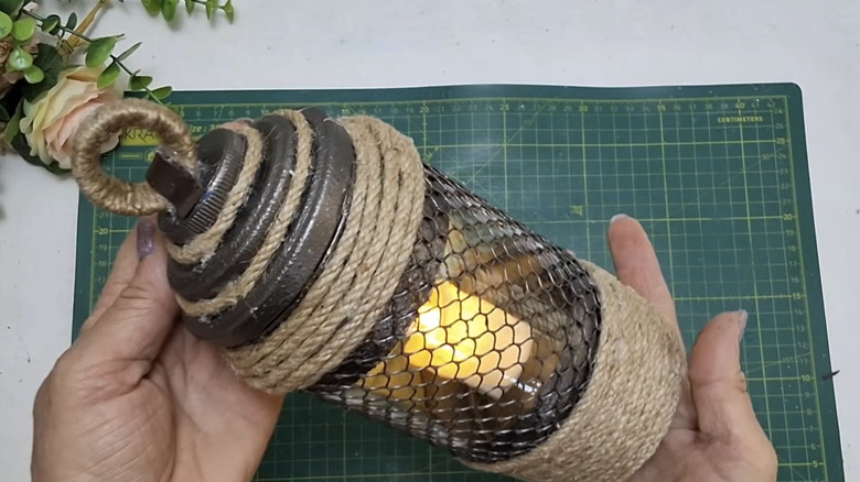 Person holding a finished DIY lantern made from a glass jar, tuna can, rope, and other crafting supplies