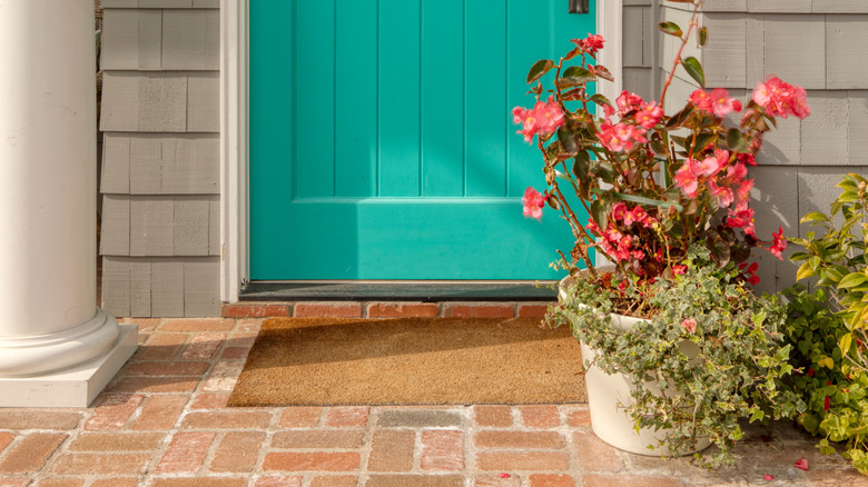Brick front porch with potted flowers and coir doormat in front of teal door.