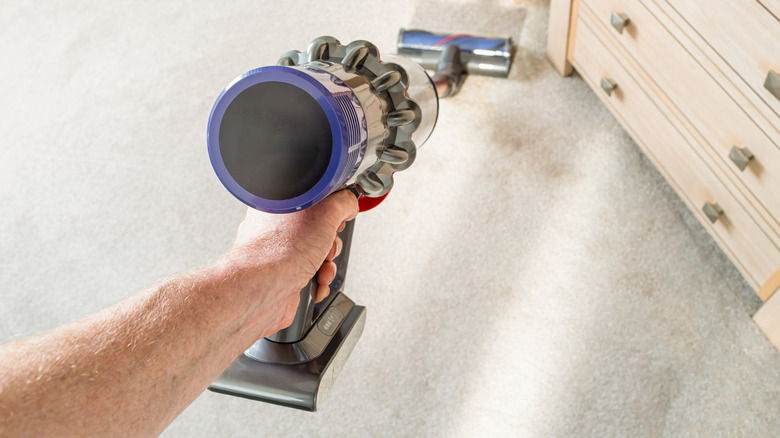 A hand holds a compact vacuum that resembles a small canister attached to a long narrow roller.