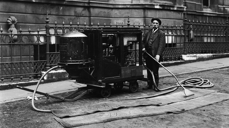 A black and white photo shows a man in a suit vacuuming a rug with a black contraption the size of a wagon.