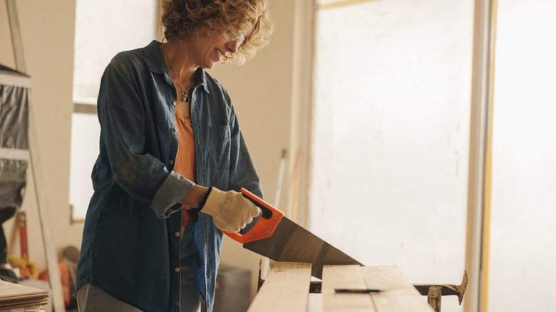 A smiling woman in a blue denim shirt using a hand saw to cut lumber.