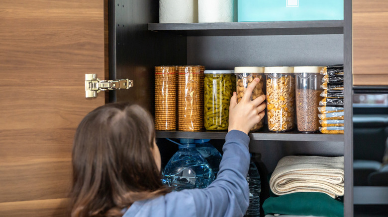 Woman is reaching into an organized pantry with shelves, holding a storage container