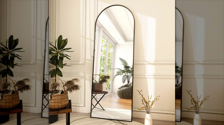 An arched full length floor mirror leans against a cream colored wall in a room filled with plants