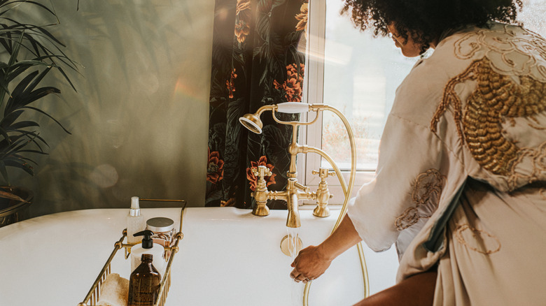 A woman holds her hands under the gold faucet of a roll top bath to access the temperature of the water as she fills the tub.