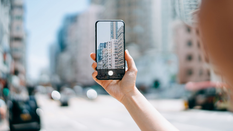A person holds a smartphone and takes a photo of a city street.