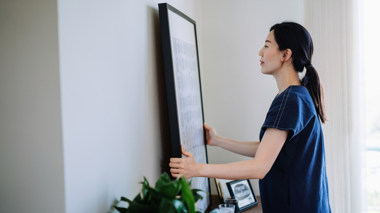 A woman holds a giant piece of framed artwork up to a wall.