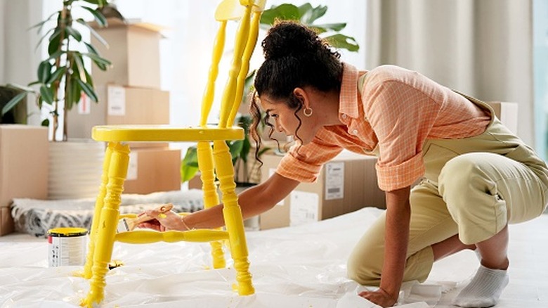 Woman painting a chair yellow in a sunny room with plants and boxes over a plastic drop cloth