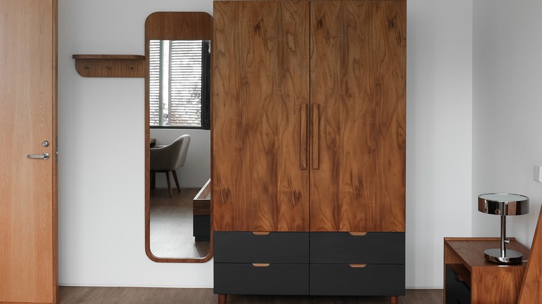 A wooden armoire with black shelves on the bottom against a wall with a mirror next to it