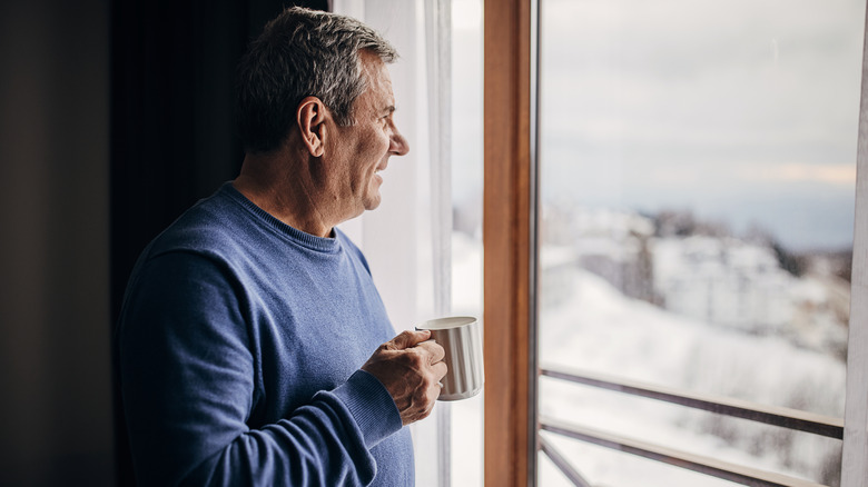 A man standing at the window admiring snow-covered trees outdoors