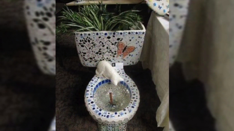 Toilet sculpture with mosaic designs and a stone cat peaking into the toilet bowl and plants in the tank