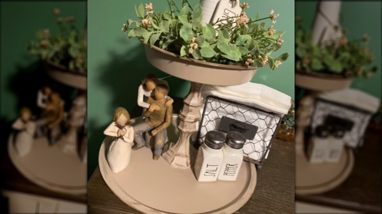 A tiered tray is covered in knick-knacks and imitation greenery and sits on a table in a green-painted room.
