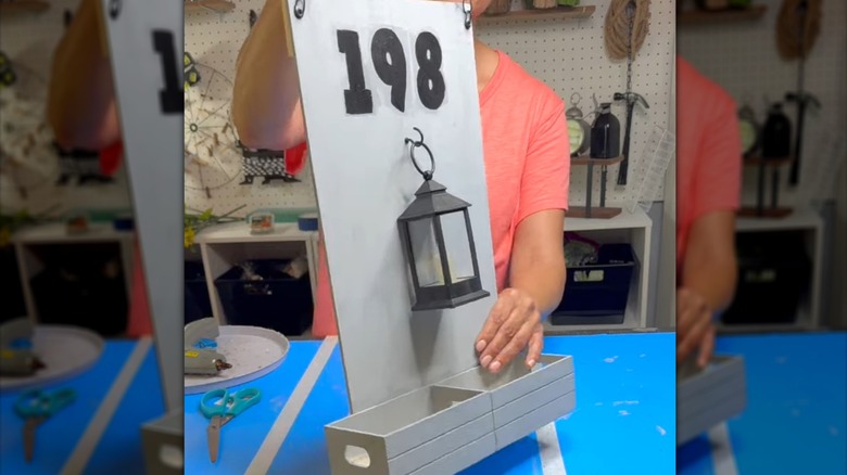 Woman holds wooden hanging house number sign made with crates and an imitation lantern