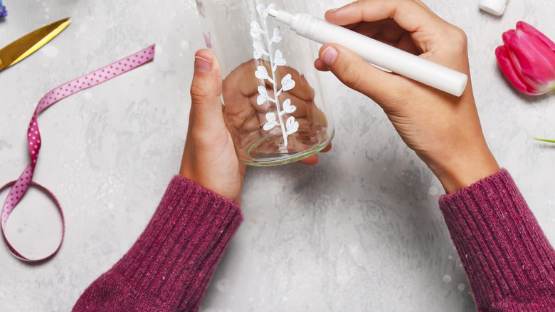 A person draws flowers on the side of a parmesan cheese container with a white permanent marker.