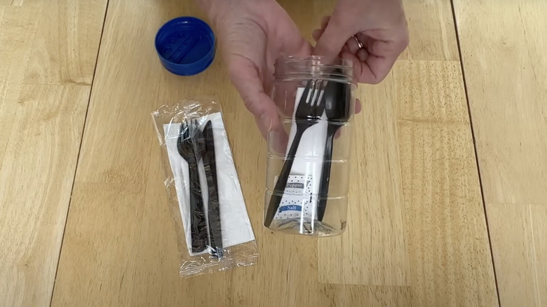 Hands holding a parmesan cheese container filled with disposable utensils and paper napkins.