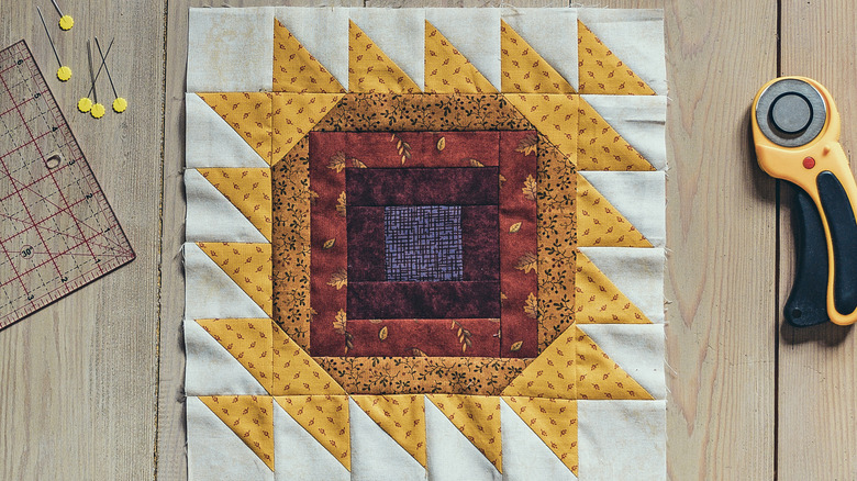 A warm-colored quilt square on a wooden surface with sewing utensils nearby.