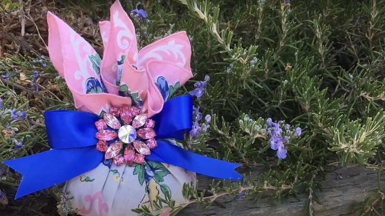 A scented drawer sachet made from a vintage hankie sits on a wall with a rosemary bush behind it.