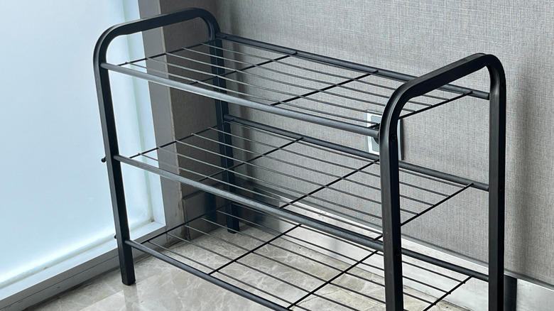 An empty, three-tiered black metal shoe rack positioned against a gray wall.
