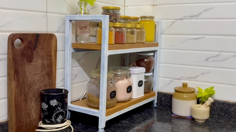 A small two-tired shoe rack used to store spices in glass jars on a kitchen counter.
