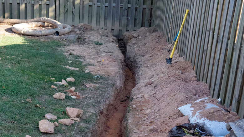 A backyard trench with a yellow trenching tool leaning against a fence.