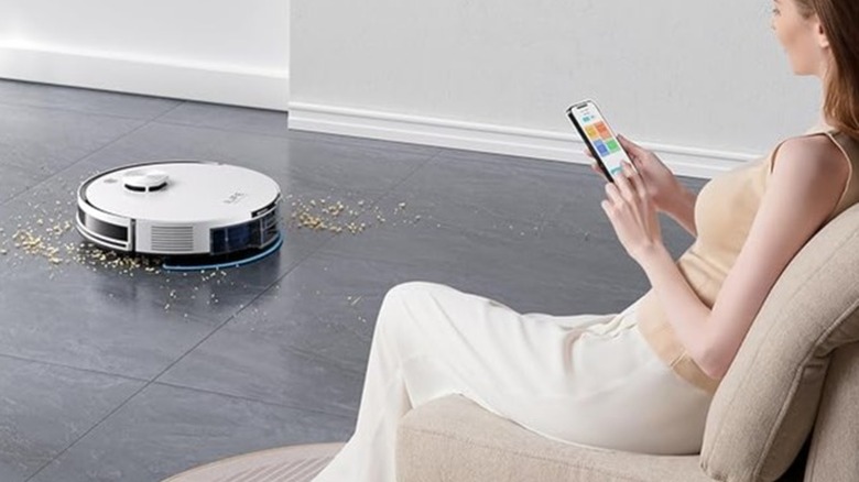 A white robot vacuum cleaning dirty floor while a woman sits nearby