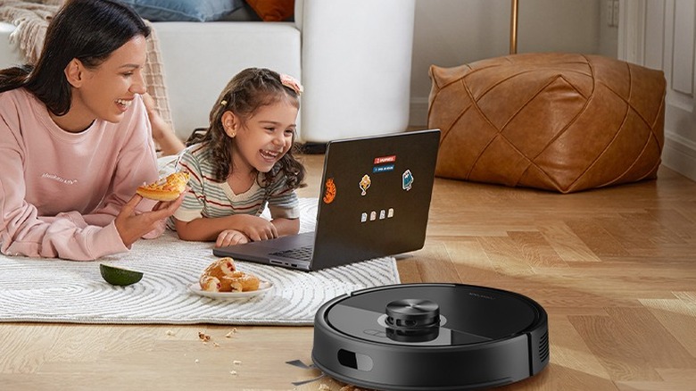 A mother and daughter snacking on carpet while a robot vacuum cleans crumbs