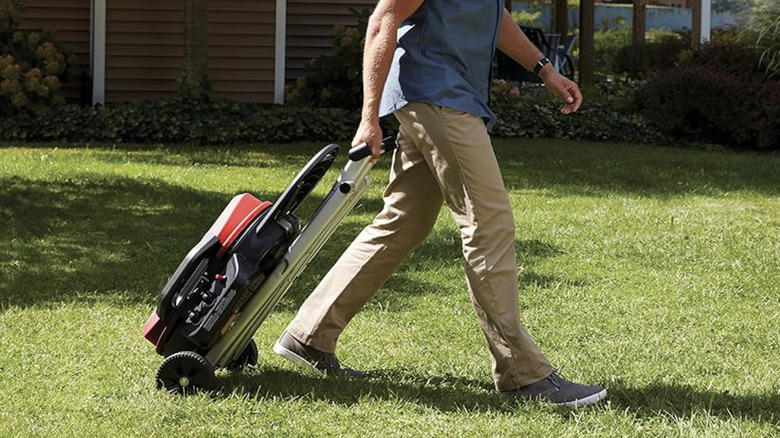 Man walking across a lawn, pulling a folded grill on wheels behind him