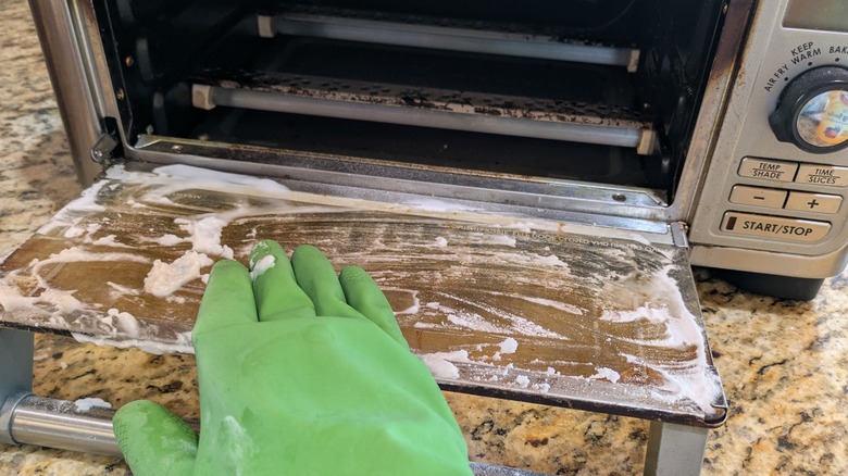 A hand in a green rubber glove wipes a cleaning paste off a toaster oven door.