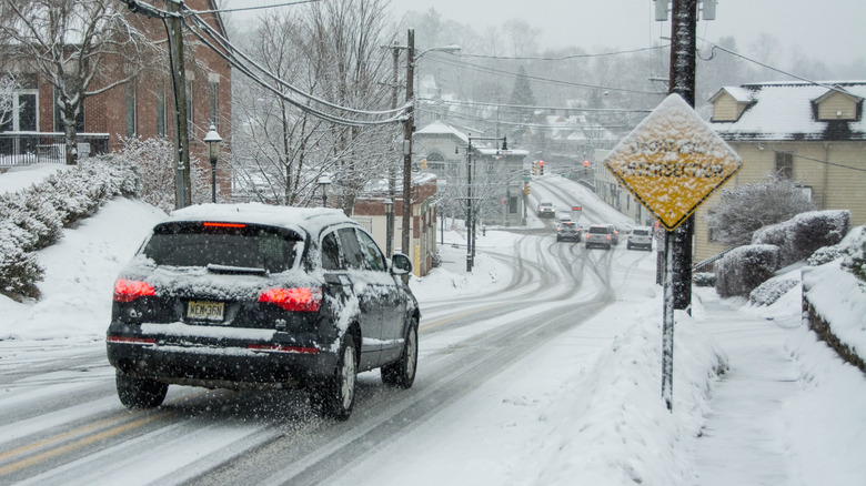Small black car driving on snow-covered street with buildings and snow-covered sign.