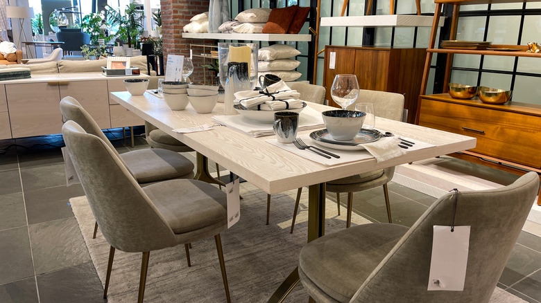 Modern wood dining room table in a West Elm store, assorted glassware including wine glasses and mugs sit on the table as it's surrounded by fabric-covered chairs